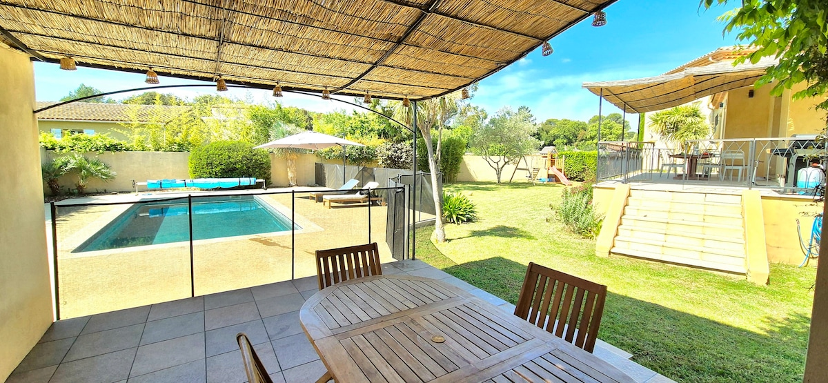 An outdoor terrace features a sturdy dining table surrounded by wooden chairs. In the background, a turquoise pool is visible, accompanied by sun loungers and a shaded seating area. Lush greenery and a play area are seen beyond the pool, contributing to the serene environment.