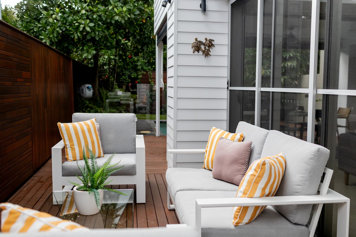 A cozy outdoor seating area is presented, featuring two armchairs and a loveseat adorned with striped cushions. A glass table sits in the center, accompanied by a small potted plant. The space is framed by wooden fencing and lush greenery, creating a serene environment.