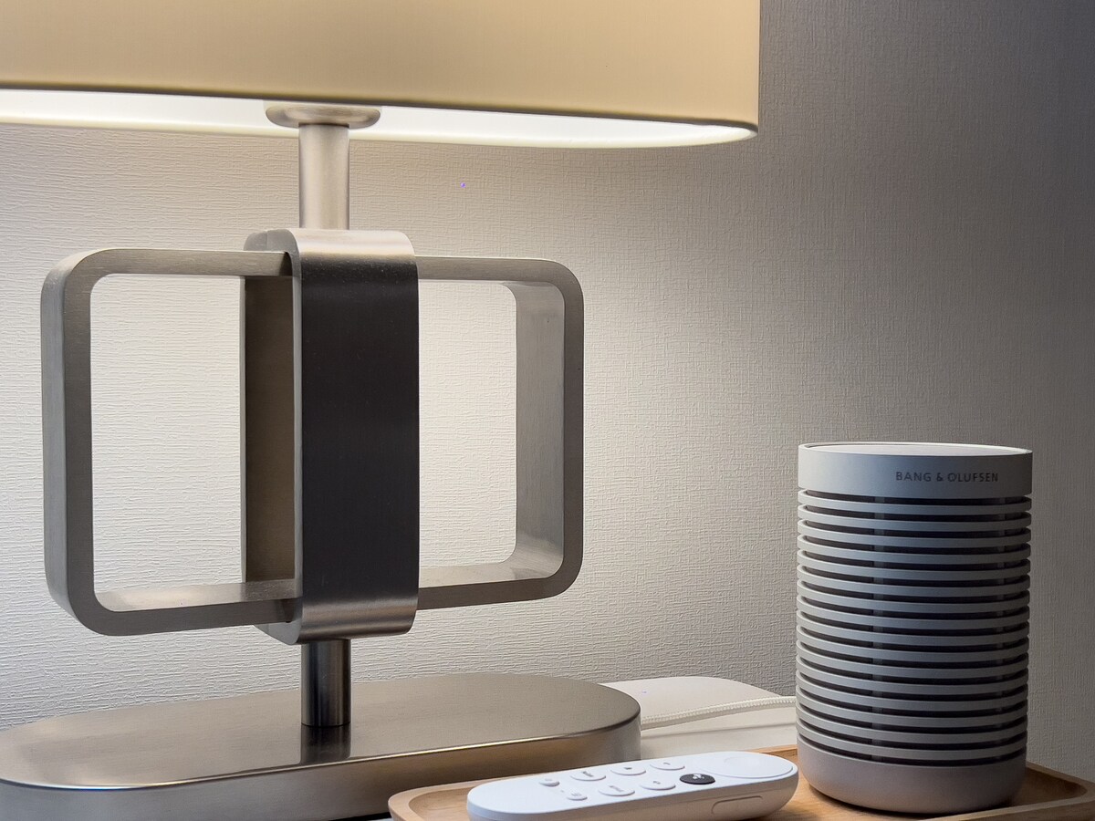 A sleek bedside table is set with a modern lamp featuring a geometric design and an ambient light shade. Next to it, a compact sound system for audio enjoyment is placed alongside a remote control for ease of use.