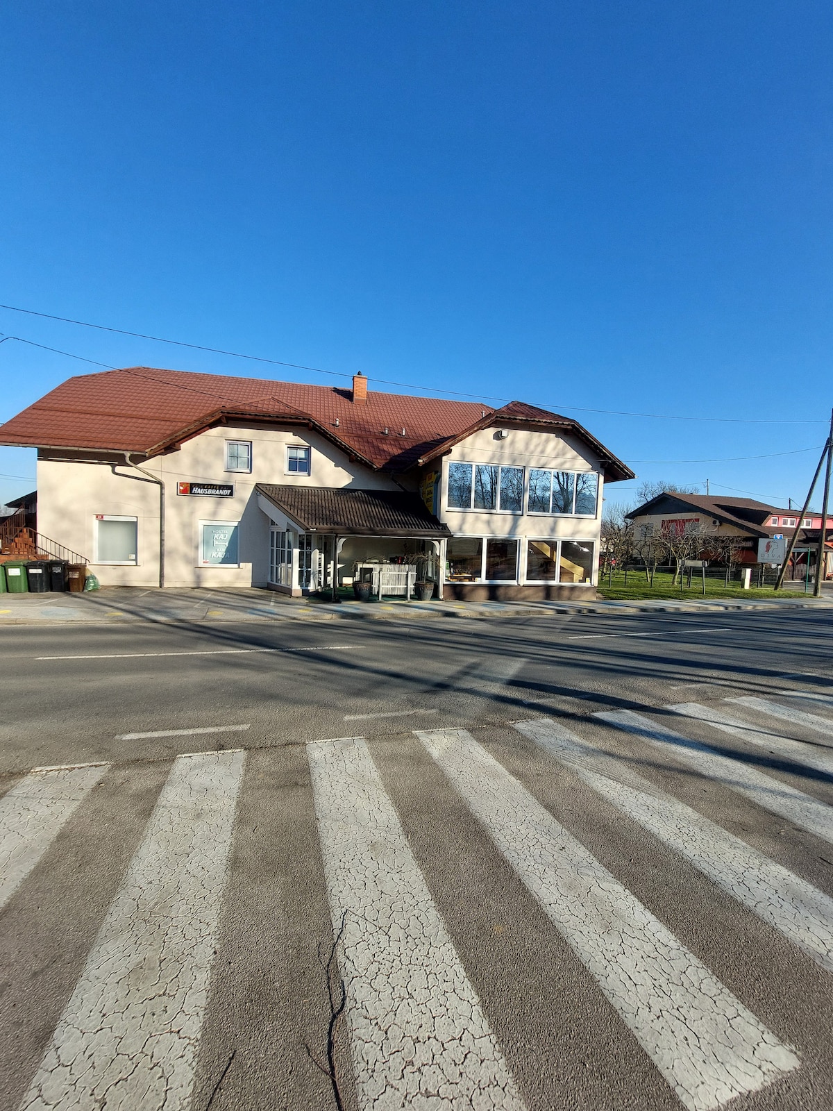 A two-story building features a sloped roof and large windows that provide natural light. The ground floor includes a covered entrance. A crosswalk in front enhances accessibility, while the surrounding area displays greenery and other structures in the distance.