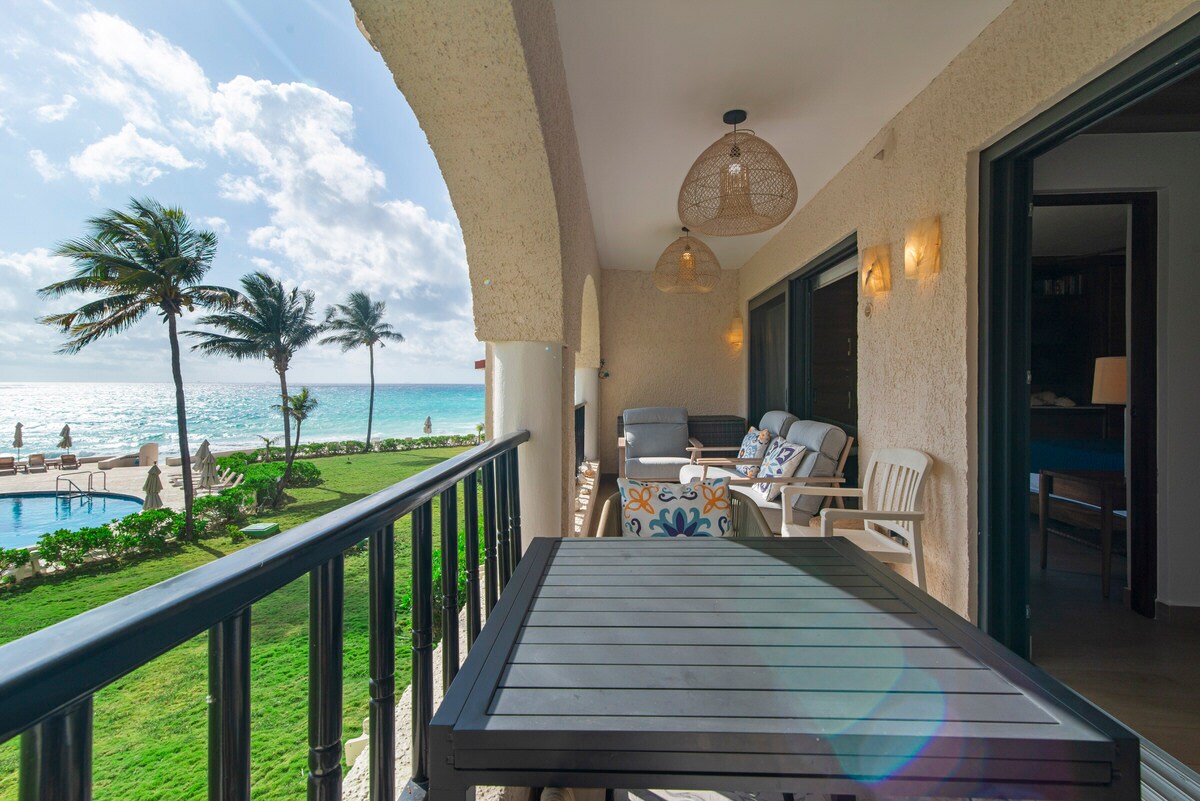 A private balcony is presented with comfortable seating arrangements, featuring a table and chairs. The view includes lush greenery and palm trees, alongside a glistening pool and the expansive ocean in the background, with soft clouds highlighted by natural light.
