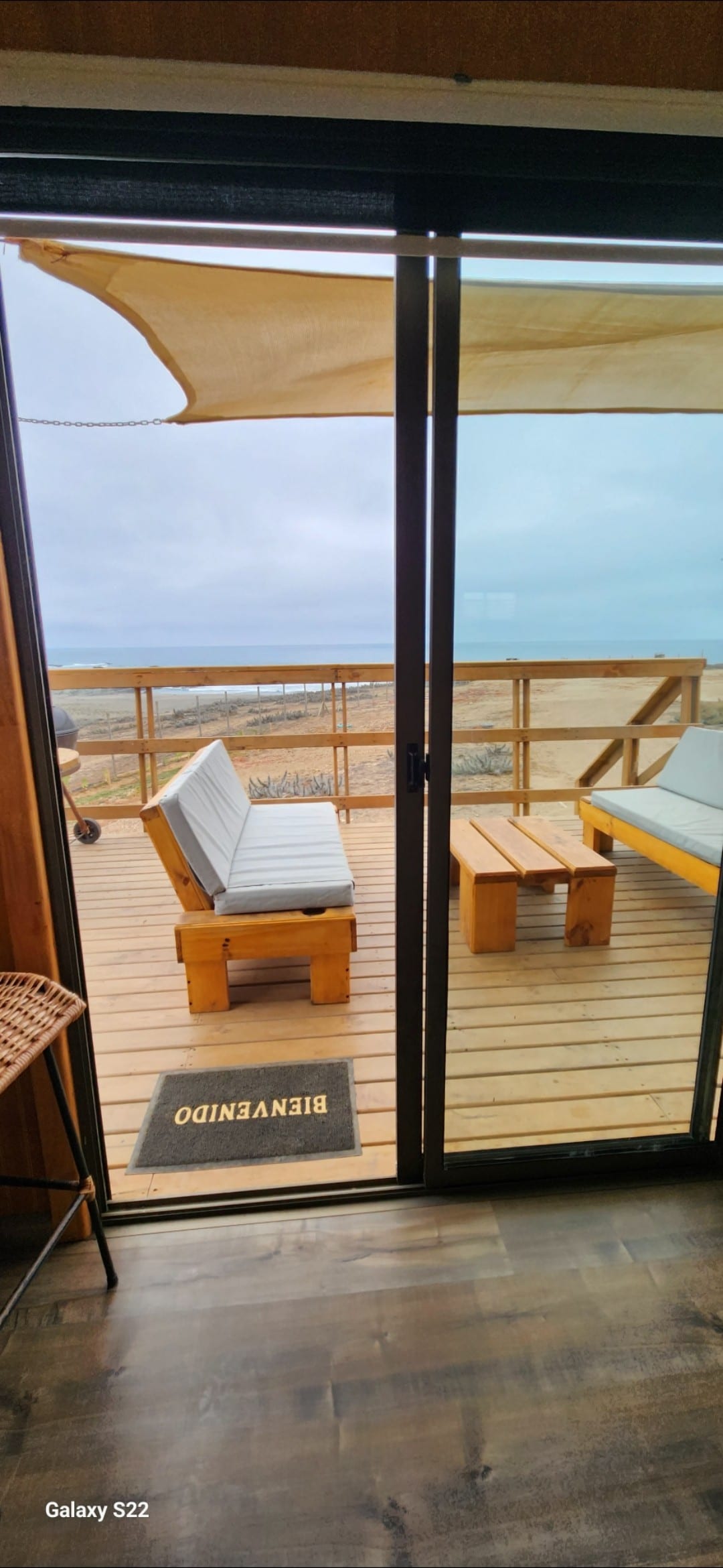 A sliding glass door introduces a spacious terrace with wooden flooring, featuring a pair of outdoor seating options and a small table. Natural light filters through, illuminating the space and providing a view of the coastline beyond.