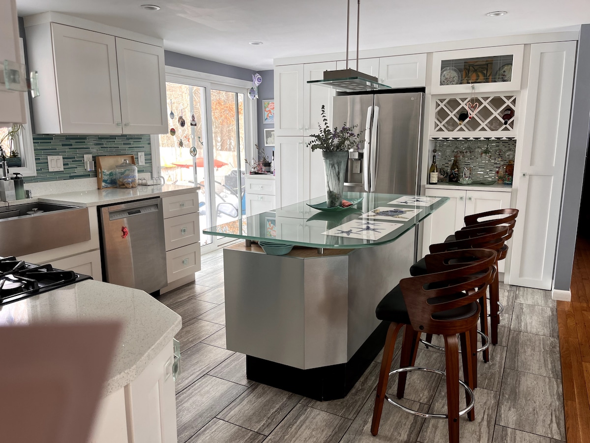 A modern kitchen features sleek white cabinetry and stainless steel appliances. An island with a glass top provides seating for four. Natural light pours in from glass doors leading to an outdoor area, enhancing the spacious layout. Backsplash tiles in varying shades complement the countertops.
