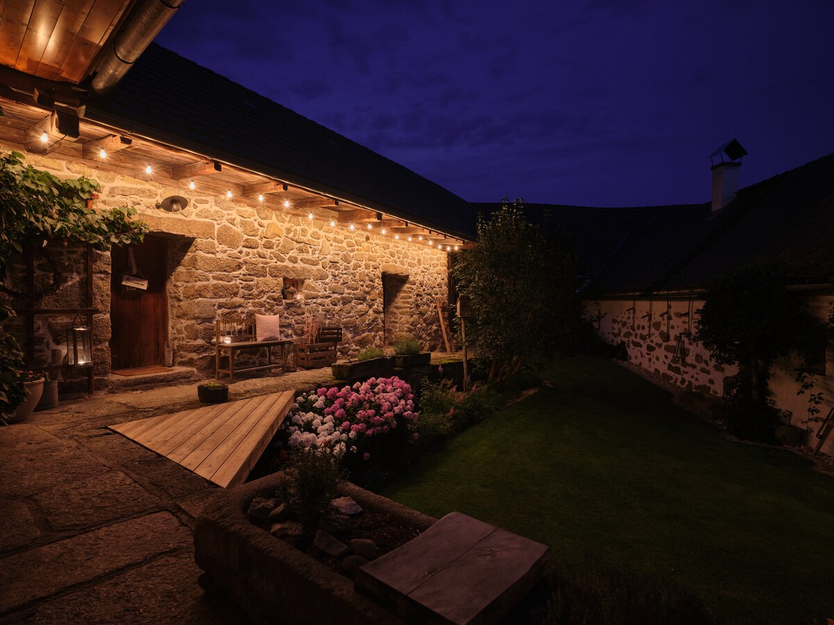 A charming stone building exudes warmth under soft outdoor lighting. A wooden deck leads towards a garden with blooming flowers and well-maintained greenery. The serene atmosphere is complemented by the visible structure reflecting the tranquil evening sky.
