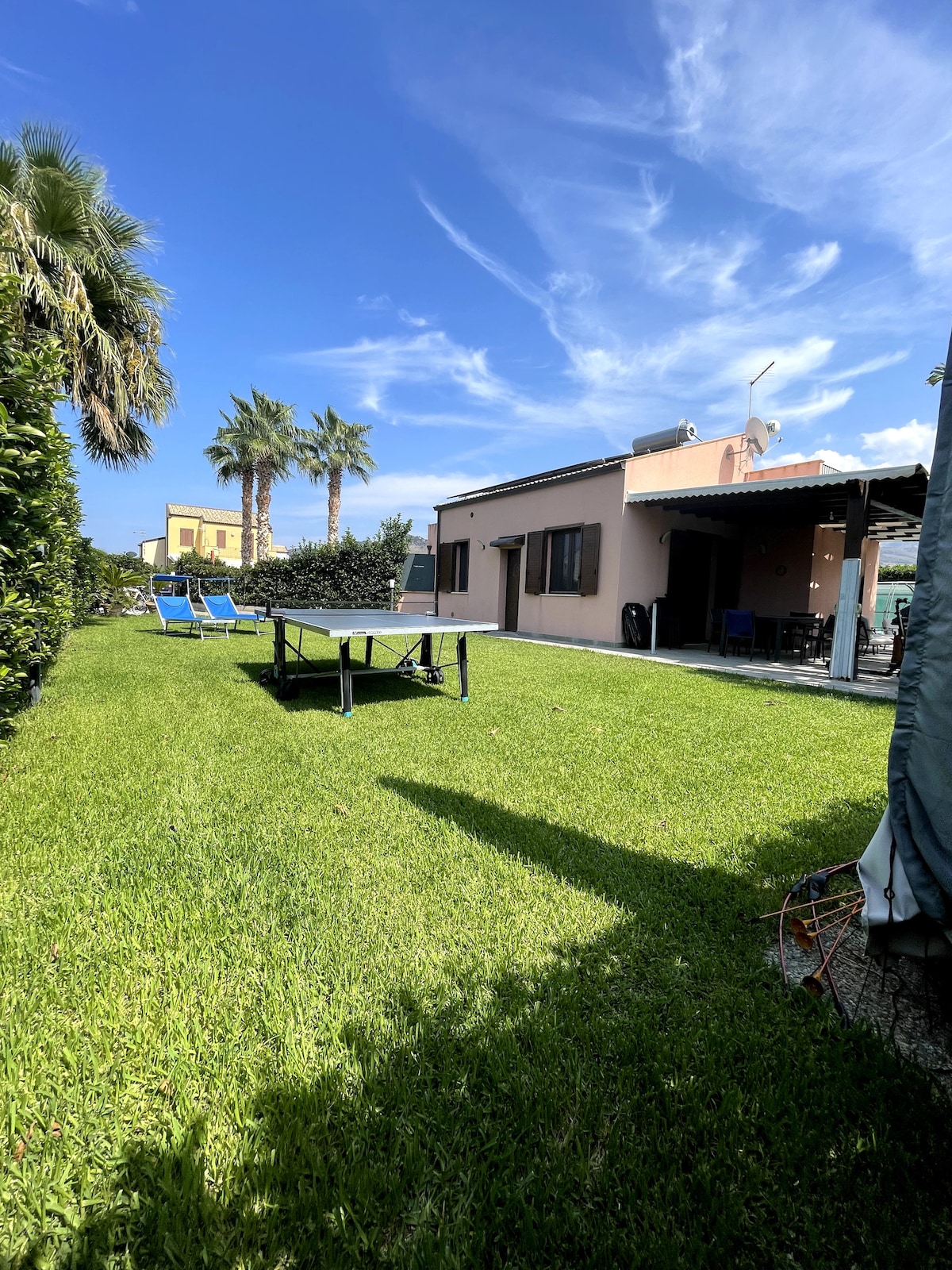 A well-maintained garden is highlighted by vibrant green grass and palm trees in the background. A table tennis setup is placed on the lawn, while a patio with outdoor seating is visible attached to the villa. The blue sky and light clouds enhance the tranquil atmosphere.