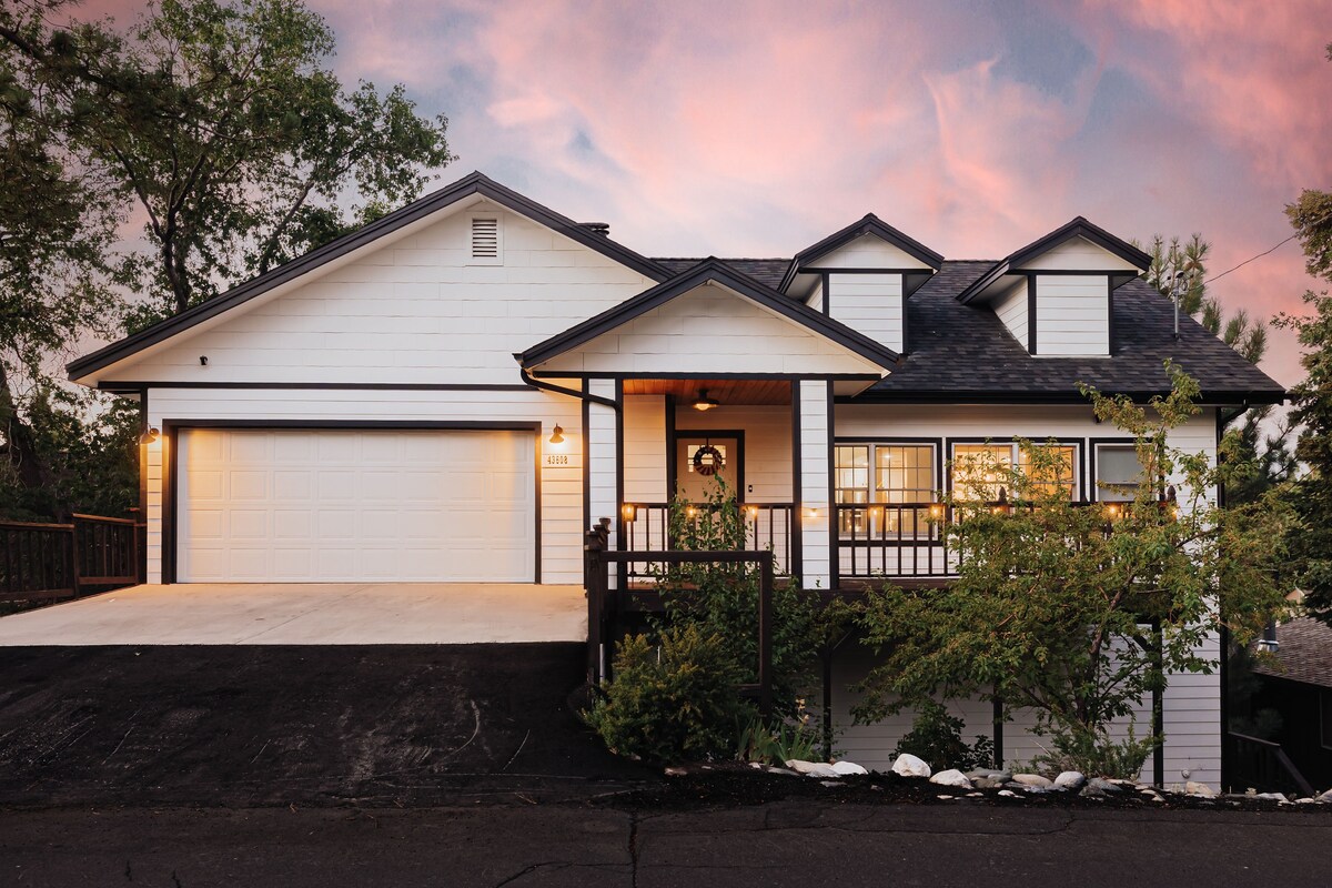 The exterior of a modern mountain home is shown, featuring a combination of white siding and dark roofing. A spacious driveway leads to a two-car garage. Lush greenery surrounds the home, and a welcoming front porch is adorned with a decorative wreath, creating a charming entry.