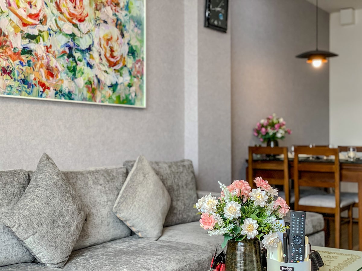 A comfortable seating area is displayed with a gray sofa adorned with several plush cushions. A floral artwork is featured prominently on the wall above. A small vase with pink and white flowers sits on a table, with a remote control and other items nearby, providing a cozy and inviting look.