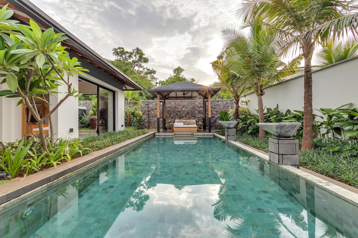 A serene pool area is framed by lush tropical plants, reflecting a clear sky above. A wooden gazebo stands nearby, offering a shaded retreat. The calm water showcases an inviting turquoise hue, enhancing the tranquil atmosphere of the outdoor space.