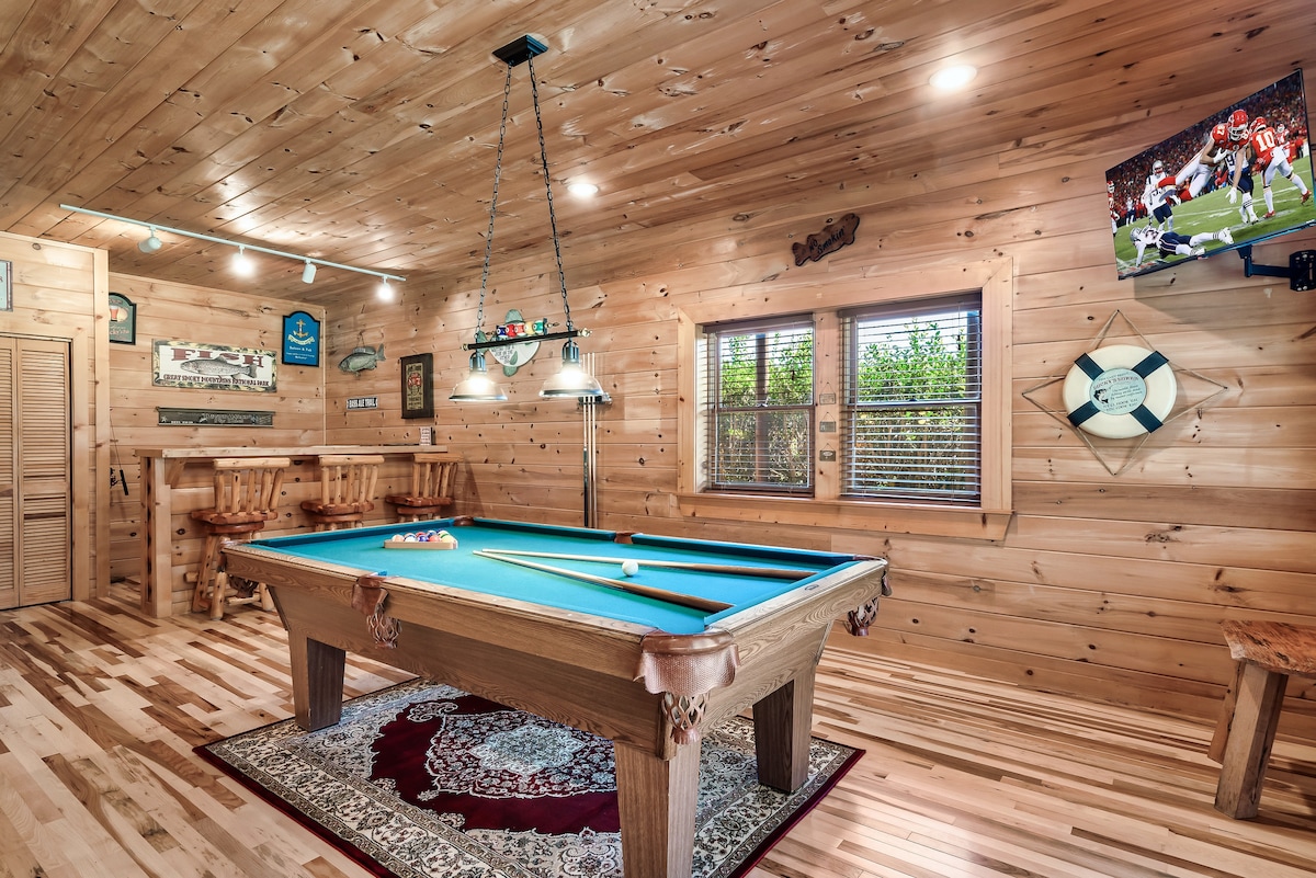 A game room with wooden walls features a billiard table with a bright blue felt surface. Hanging lights illuminate the area. A flat-screen TV is mounted on the wall, displaying a sports game. Nearby, a bar with stools provides additional seating options.
