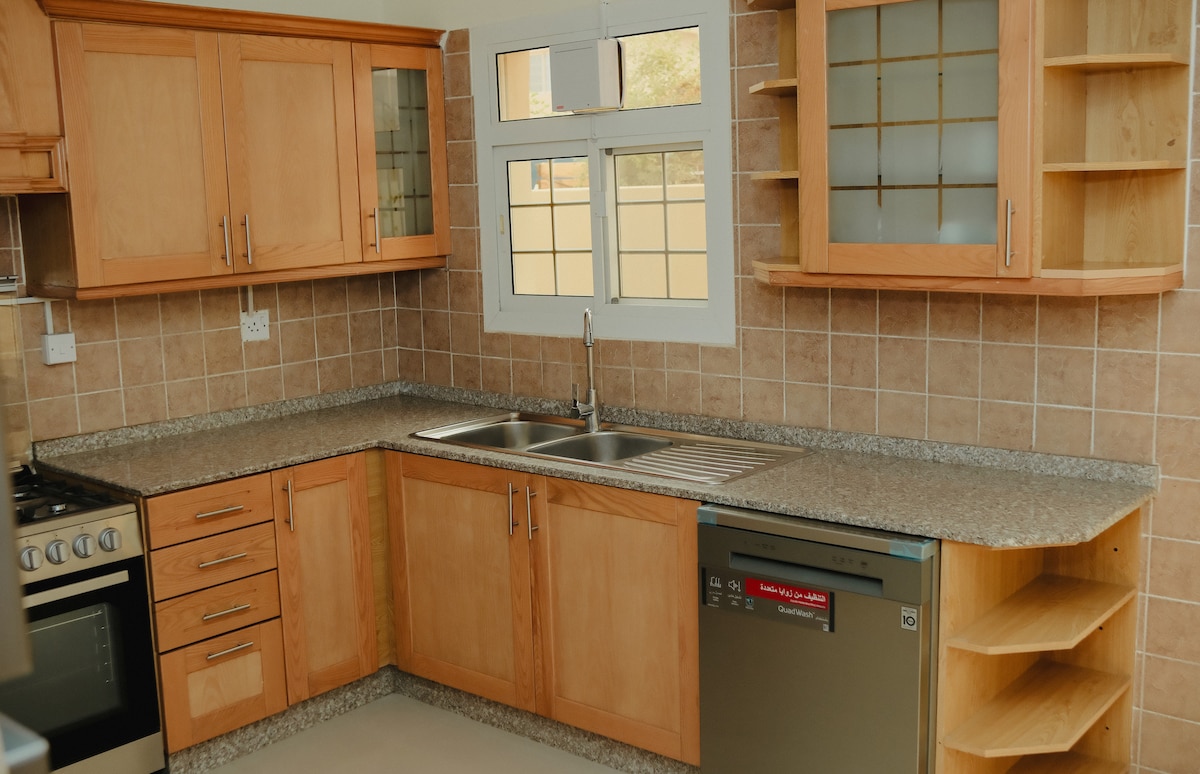 The kitchen features light wood cabinetry with integrated shelving and a spacious granite countertop. A double sink is positioned beneath a window, allowing natural light to enter. Modern appliances include a stove and dishwasher, all set against a tiled backsplash.