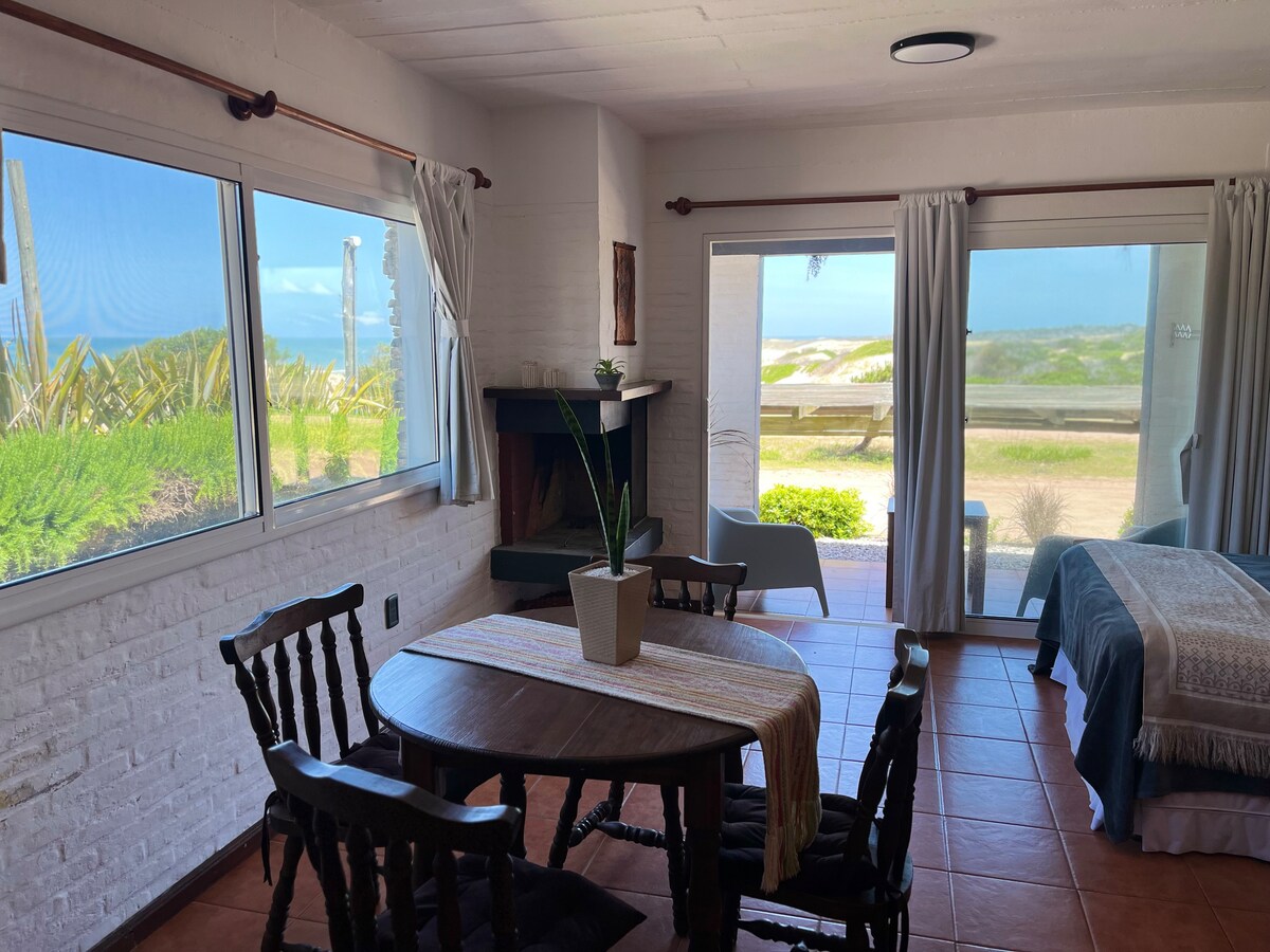 A cozy living space features large windows that frame views of the natural landscape and the ocean. A wooden dining table with chairs sits near a compact kitchenette, while a fireplace adds a homey touch. The room is well-lit with neutral tones.