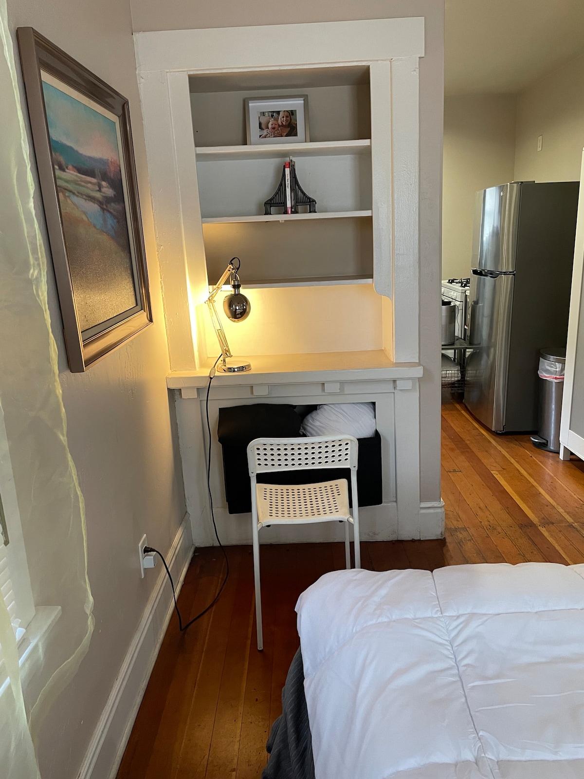 A cozy workspace is highlighted by a wall-mounted lamp and a simple chair beside a small shelf displaying a framed image. The warm wood floor extends into the background, where a modern kitchen refrigerator is visible.