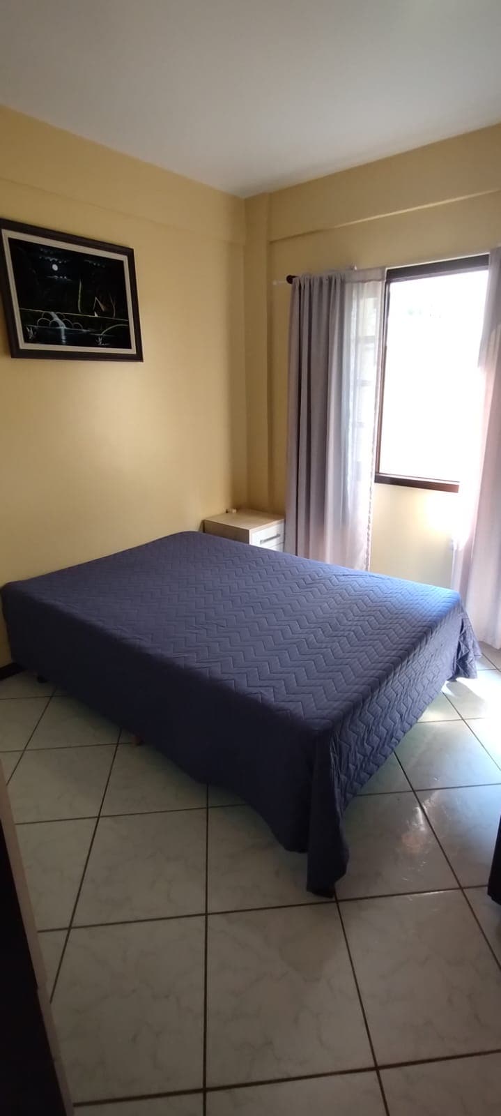 A well-lit bedroom features a spacious bed with a blue quilt, complemented by tiled flooring. Natural light streams through a window dressed with sheer curtains, while a framed artwork adorns the wall above. A bedside table sits beside the bed, contributing to the cozy ambiance.