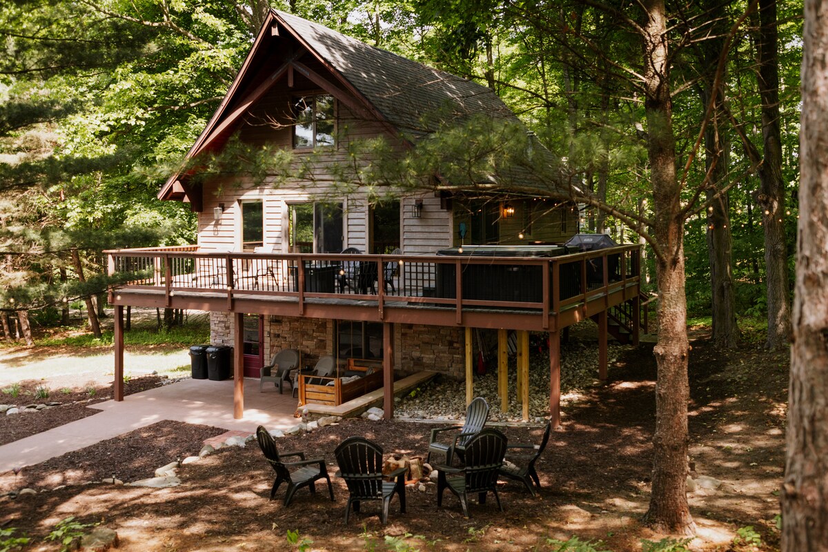 The chalet is situated on a wooded lot, featuring a large deck with outdoor seating. Below the deck, a fire pit area includes several black chairs, surrounded by trees, providing a natural and serene atmosphere.