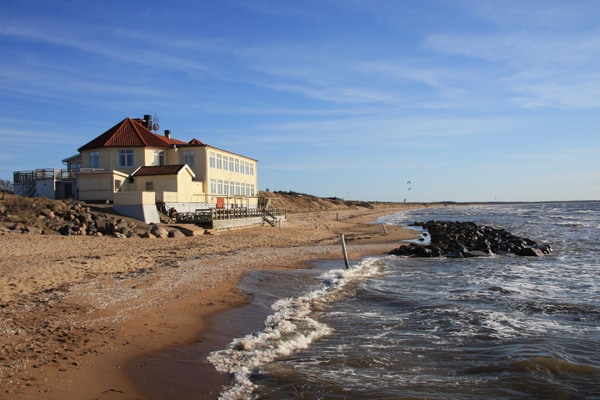 A beachside restaurant is situated near the shoreline, featuring large windows and a distinctive red roof. The sandy beach stretches along the coast, with gentle waves lapping at the shore. Rocky formations are visible in the water, contributing to the natural scenery.