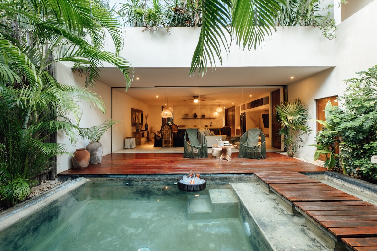 A lush outdoor space features a private pool surrounded by greenery. Wooden decking extends from the pool to an interior living area, where large glass doors create an open atmosphere. Comfortable seating is visible, inviting relaxation in a tranquil environment.