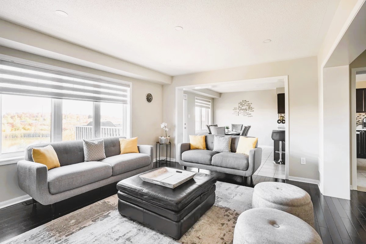 An elegant living area features two plush sofas with accent pillows, a coffee table, and a modern rug. Large windows provide ample natural light and showcase views of the surrounding landscape. A dining area is visible in the background, enhancing the open concept feel.