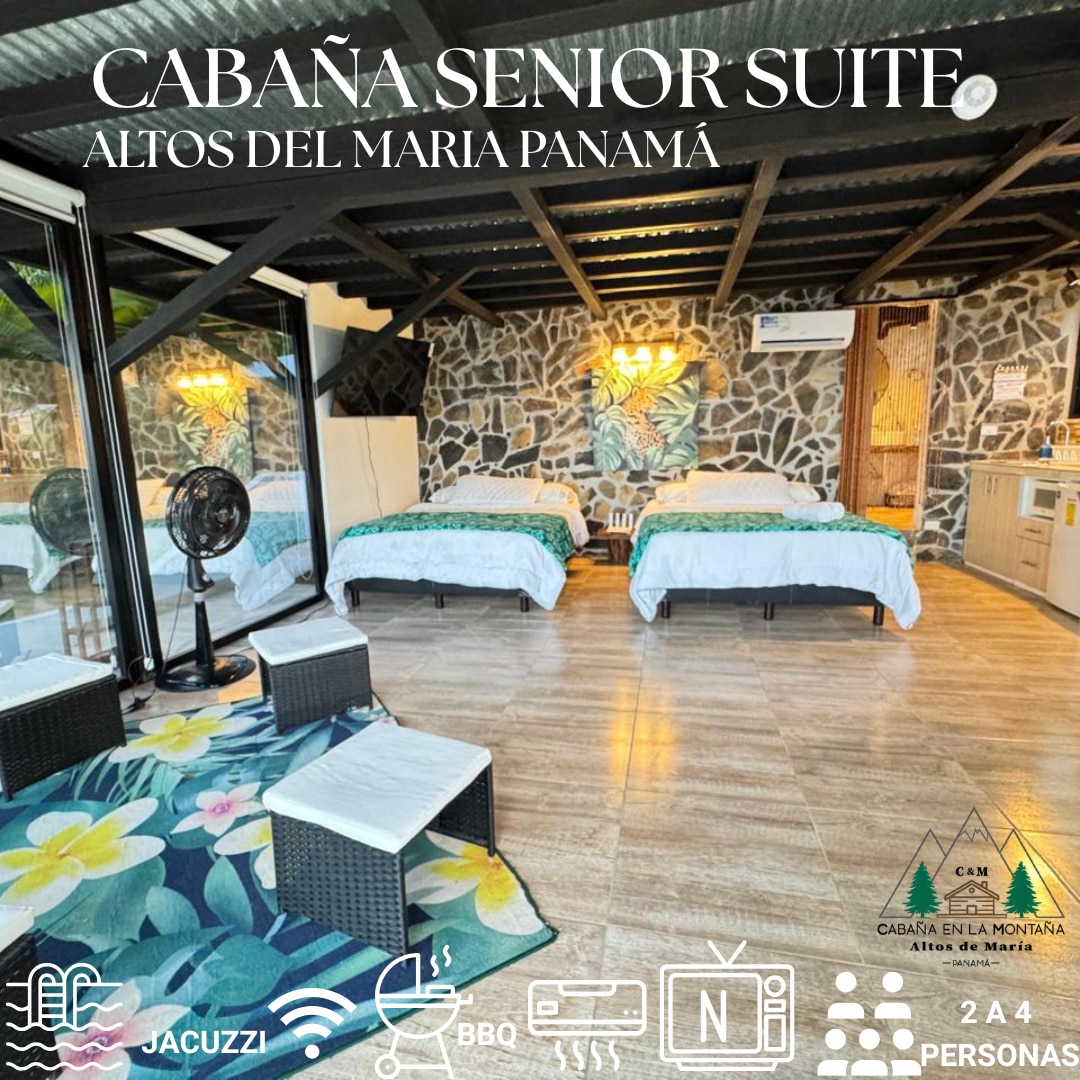 The spacious cabin features two large beds, set against a stone wall backdrop. Large glass doors provide natural light and offer views of the surrounding landscape. A cozy seating area is present, complemented by floral decorations on the floor.