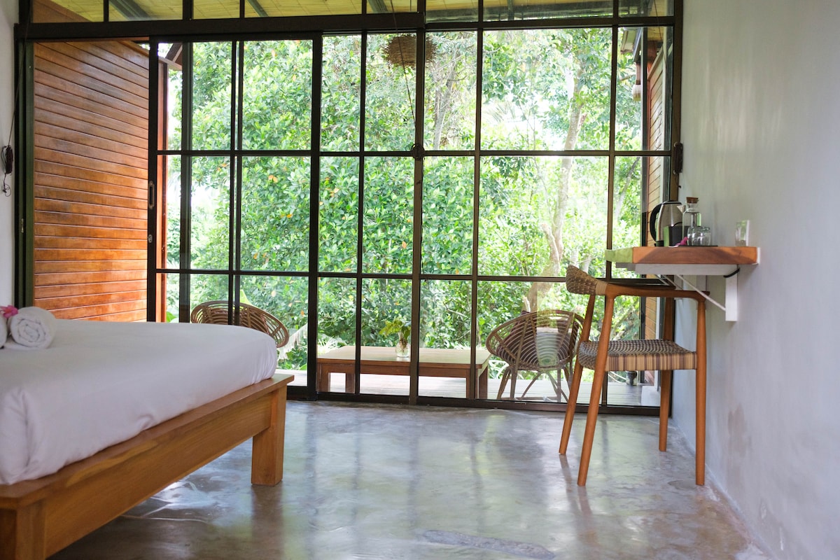 A light-filled room showcases a spacious bed with crisp linens and a calming atmosphere. Large glass doors provide unobstructed views of the lush greenery outside, while a small wooden desk and two wicker chairs are positioned for relaxation or work.