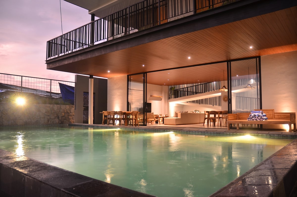 A modern villa features a private heated swimming pool surrounded by a spacious outdoor deck. Glass doors lead into a beautifully lit living area, with dining tables visible. Subtle lighting enhances the tranquil atmosphere, while the backdrop of mountains adds depth to the scene.