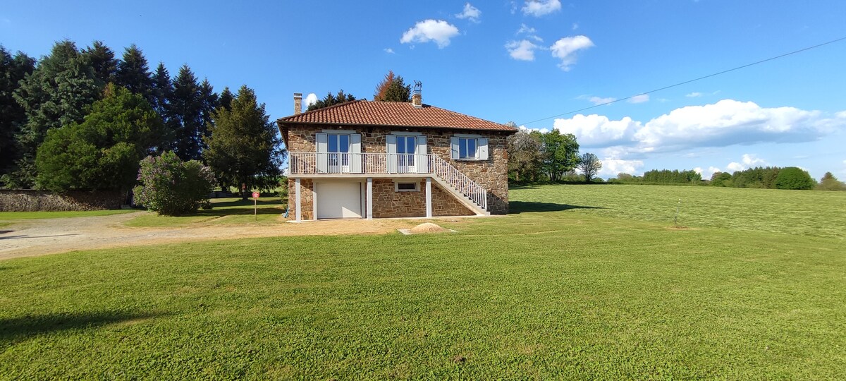A stone house with a sloped roof is situated in a grassy area, surrounded by trees. Large windows reflect the blue sky, while a staircase leads to the entrance. An expansive lawn extends in front, offering an open view of the natural landscape.