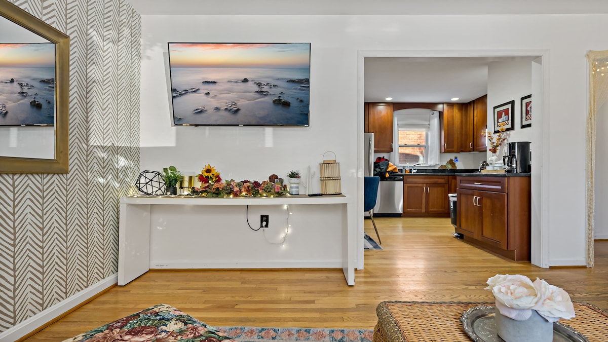 A living area is depicted with a flat-screen TV mounted above a decorative shelf adorned with plants and flowers. The room features wooden floors and a large mirror reflecting light. The kitchen is visible in the background, showcasing wooden cabinetry and modern appliances.