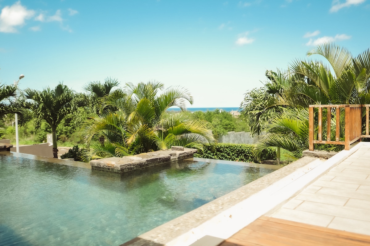 An expansive infinity pool reflects the bright blue sky, bordered by lush palm trees and tropical foliage. The tranquil water offers a seamless view of the horizon, enhancing the serene outdoor setting. A wooden deck leads to the pool, creating an inviting space for relaxation.