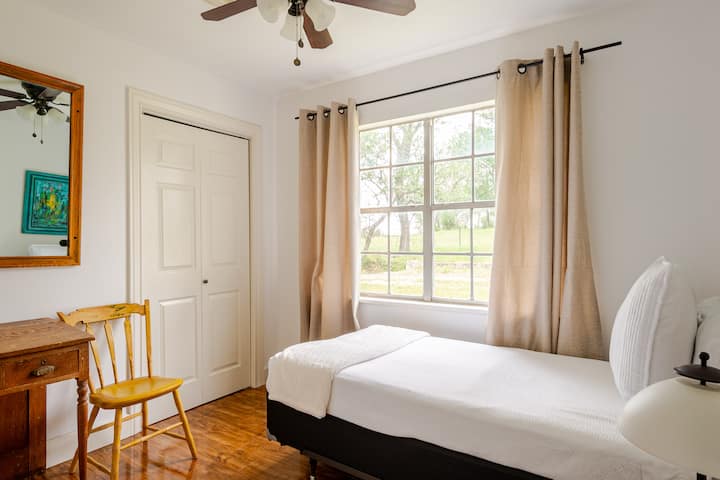 This fourth bedroom showcases a single-size bed and is beautifully decorated with bright white and pops of color. It includes a work desk and a large window overlooking the back of the property, where you may spot an Oreo cow grazing nearby.