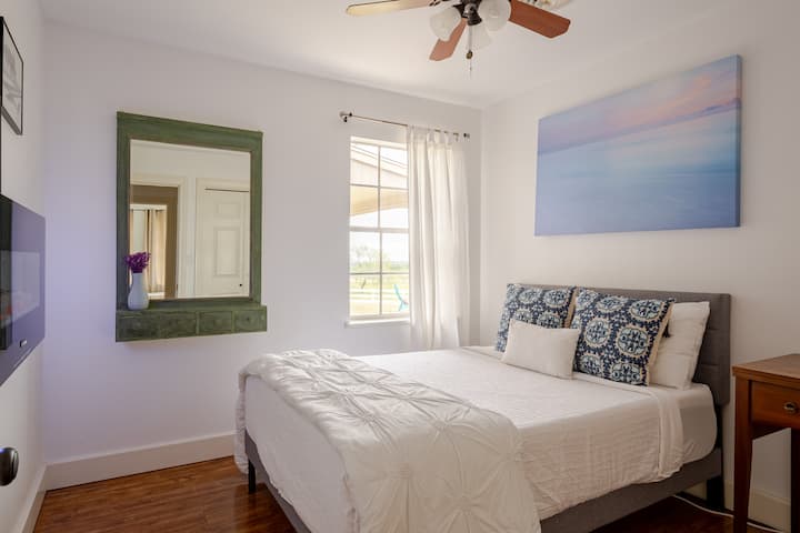 Bedroom #3 features a cozy bed, a wall-mounted mirror, and a stunning picture of a blue ocean and pink sky sunset that sometimes mirrors the actual sunsets visible from this window.