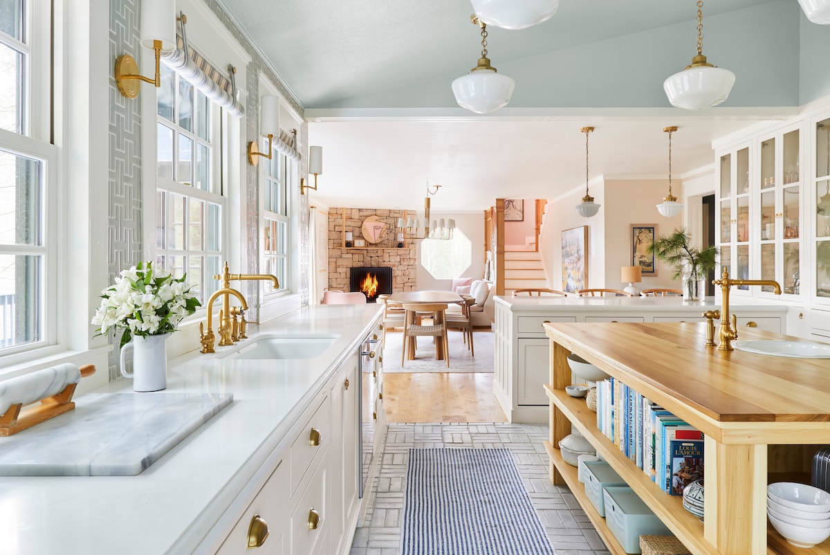 A spacious kitchen is highlighted by white cabinetry and elegant brass fixtures. A large island with a wooden countertop provides additional storage and workspace. Sunlight streams through multiple windows, illuminating the room and creating a warm atmosphere. A cozy living area with a stone fireplace is visible in the background.