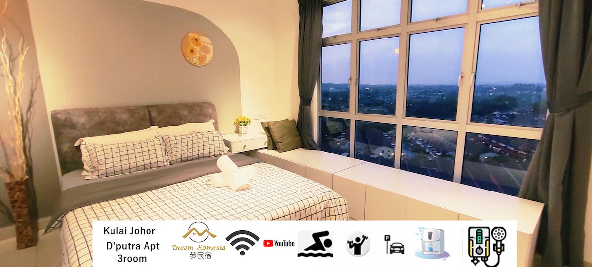 A spacious bedroom is softly illuminated by natural light from large windows, offering views of the surrounding area. The room features a queen bed with a checkered bedspread and a bedside table. A cozy ambiance is created with neutral-toned walls and light curtains.