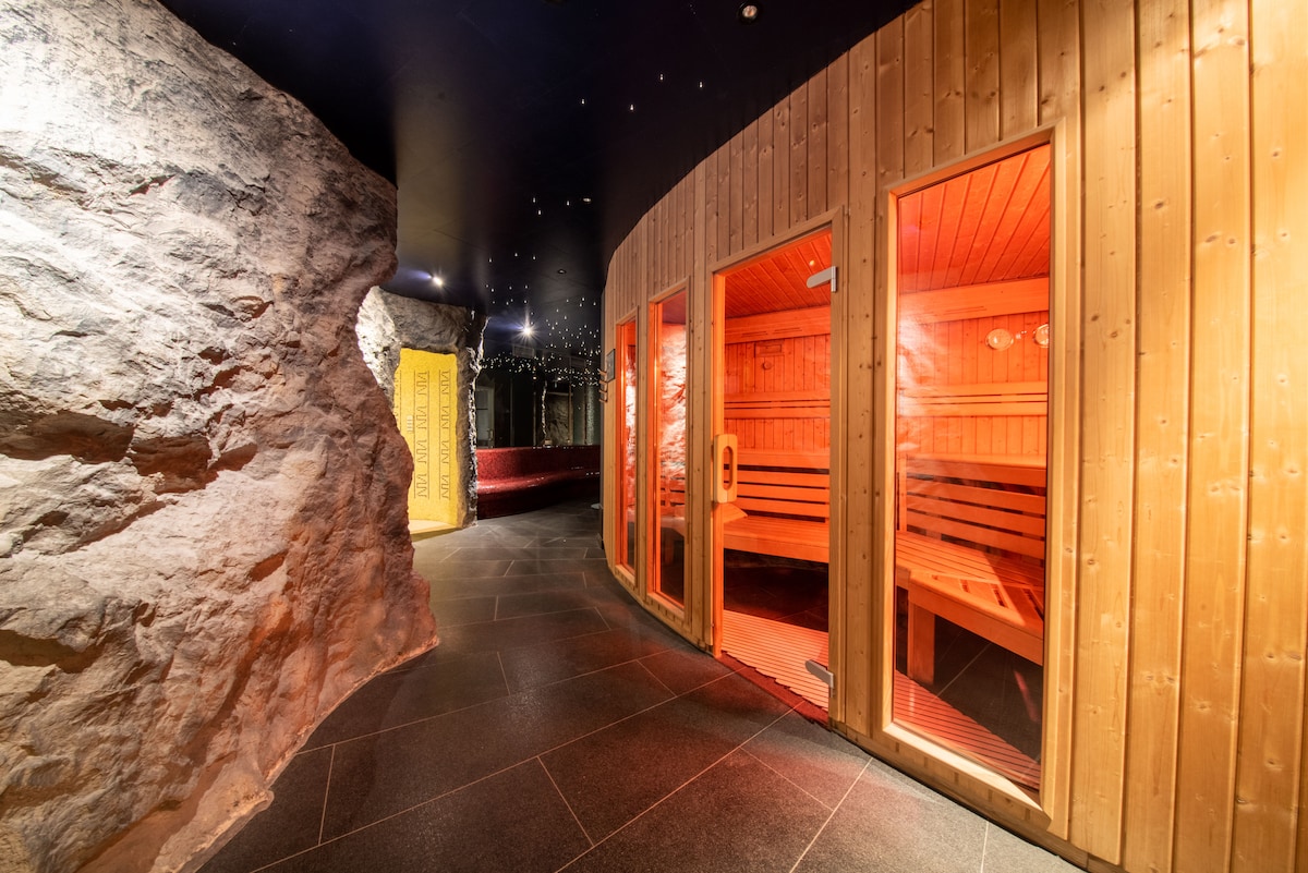 A serene sauna area features wooden walls and benches, illuminated by warm lighting. The entrance to the sauna is highlighted by glass doors, and natural stone accents create a calming atmosphere. Soft, dim lighting enhances the overall ambiance, inviting relaxation.
