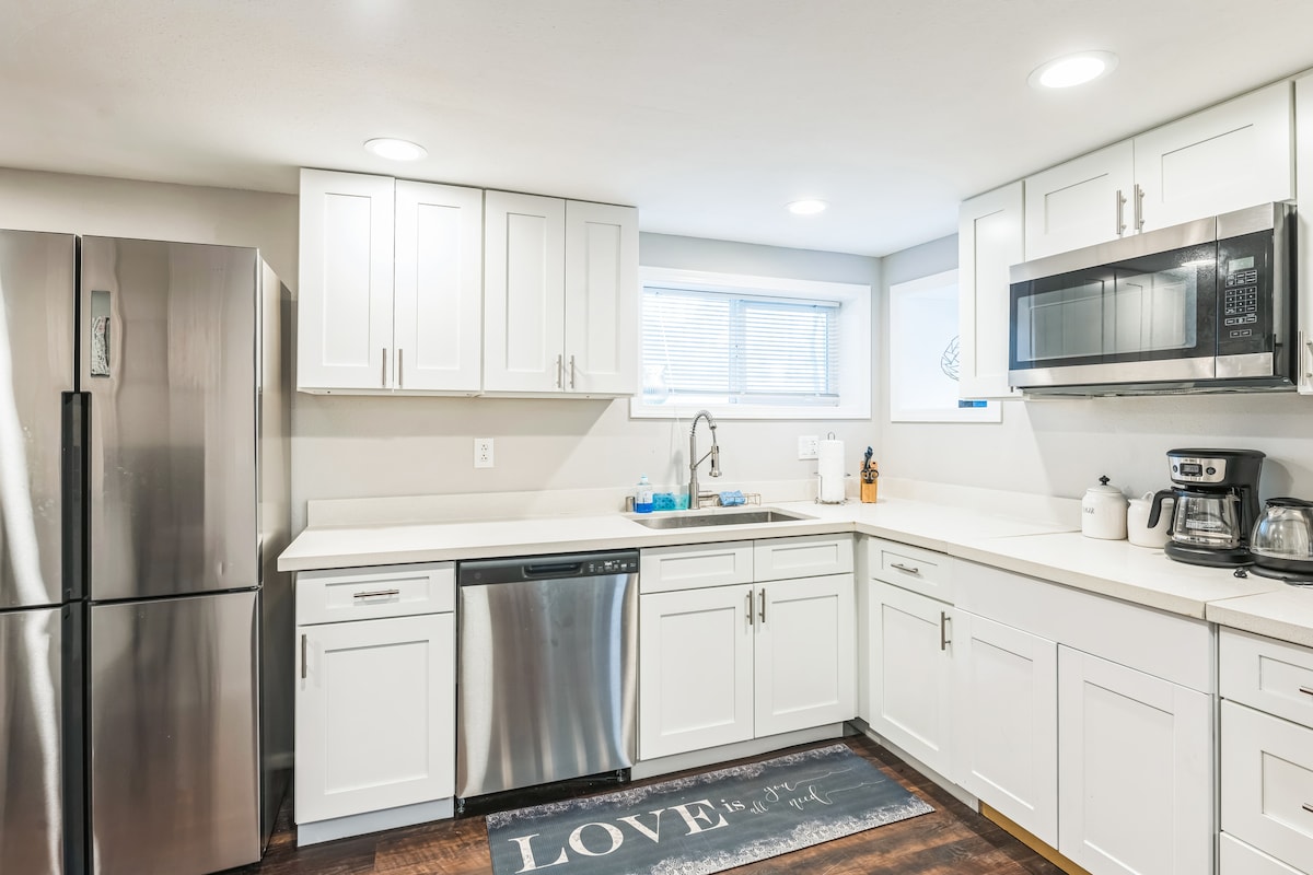 The kitchen features modern white cabinetry, stainless steel appliances, and a large countertop area. Natural light enters through a window, illuminating the space. A coffee maker and kettle are positioned on the countertop, offering convenience for meal preparations.