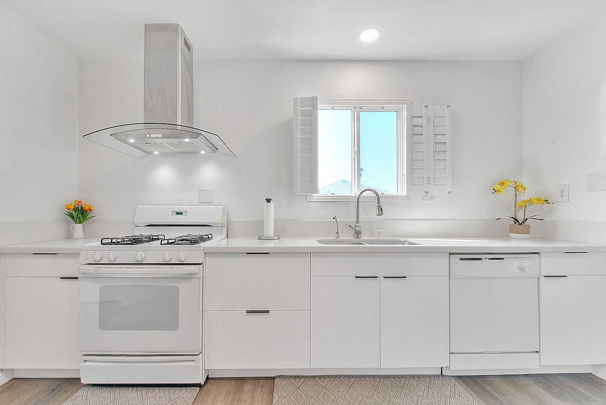 A modern kitchen features sleek white cabinetry and a stainless steel gas stove. Natural light enters through a window adorned with shutters, and a potted plant adds a touch of color to the countertop. Essential kitchen appliances, including a dishwasher, are neatly integrated into the design.