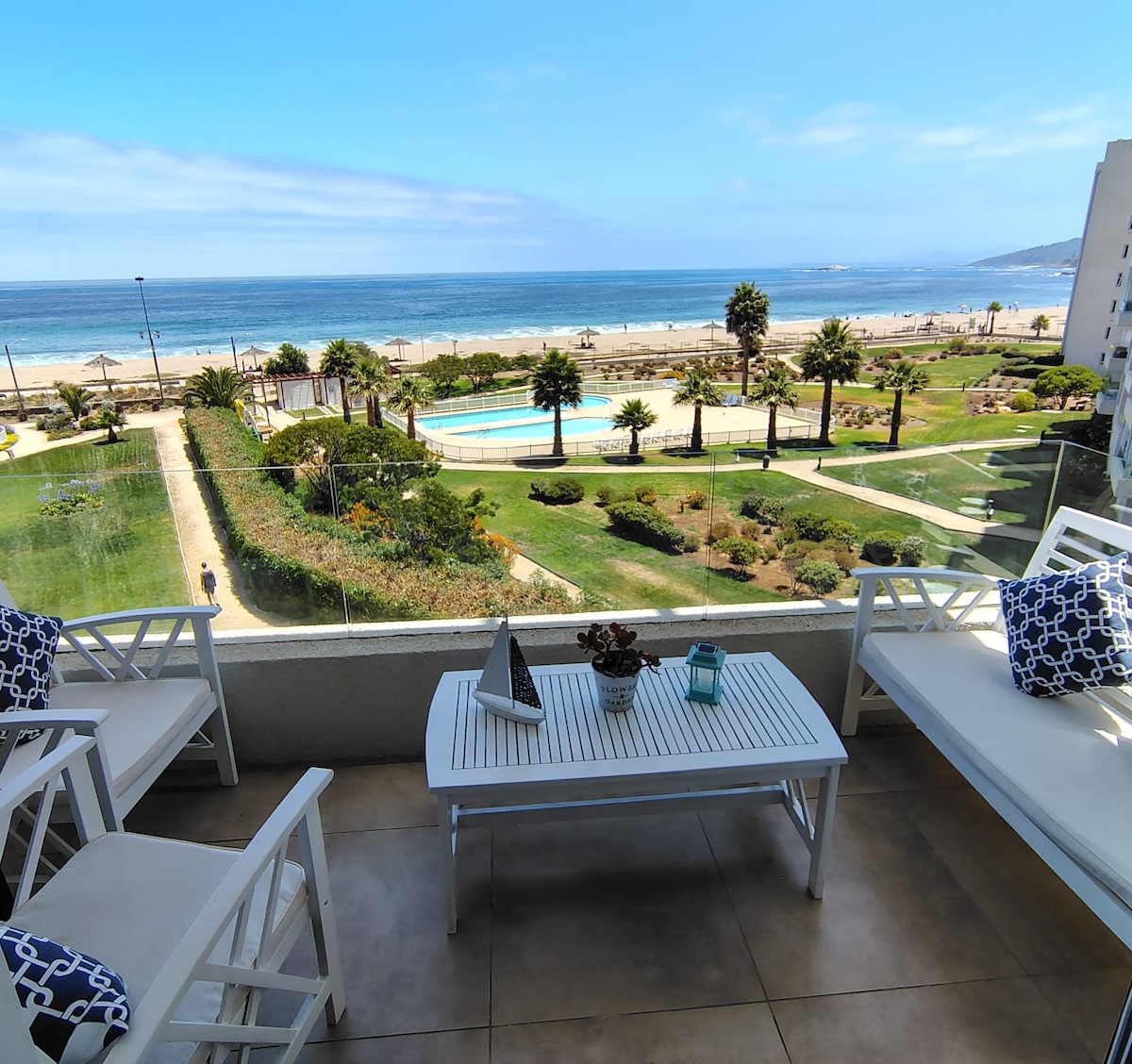 A terrace is shown with white outdoor furniture and decorative cushions, positioned to overlook a pool and expansive green lawn. The ocean can be seen in the background, complemented by a clear blue sky, creating a serene coastal setting.
