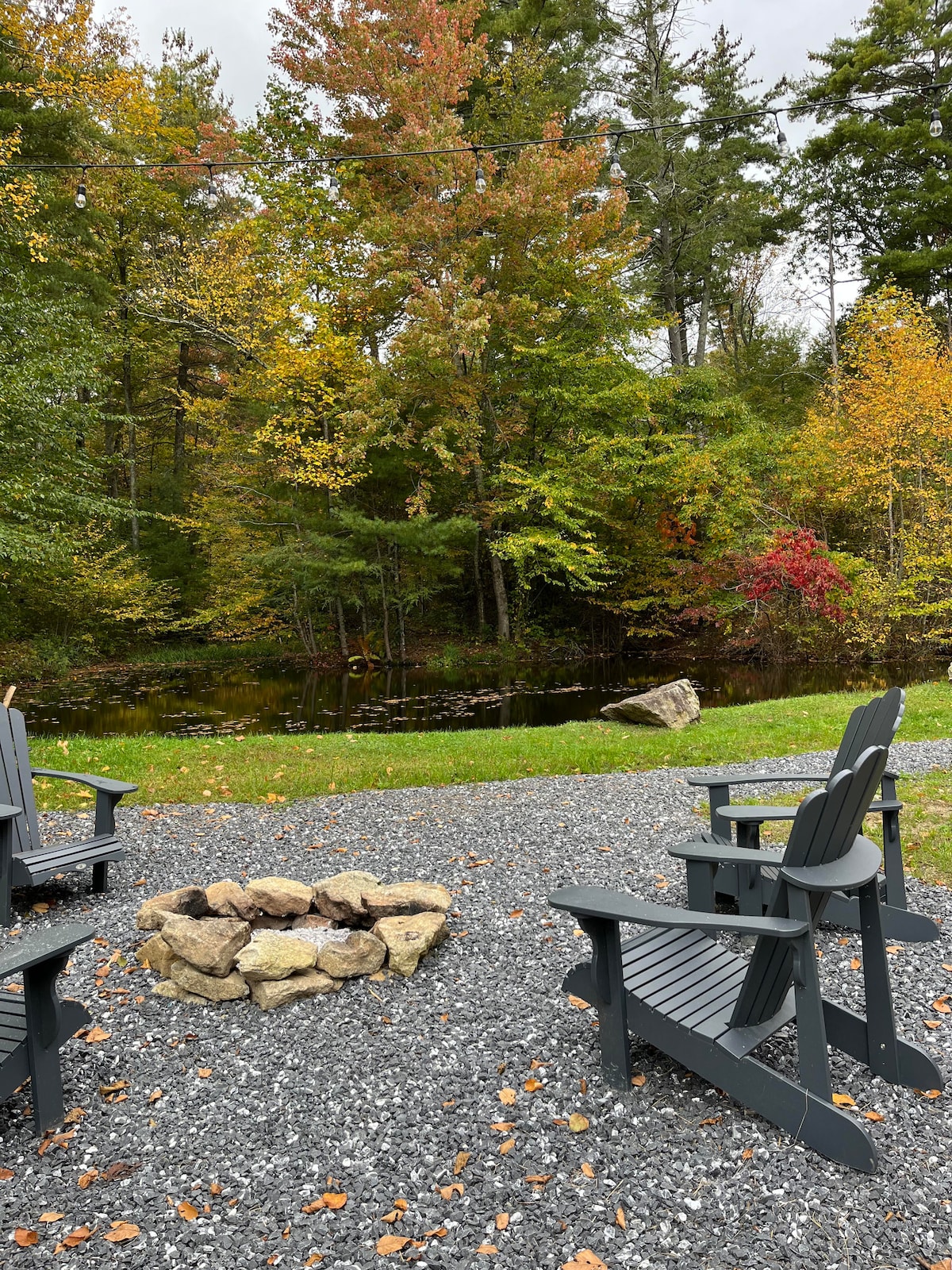 A fire pit is surrounded by several dark wood chairs, situated on a gravel area. In the background, colorful trees reflect autumn hues, and a peaceful pond is visible among the greenery.