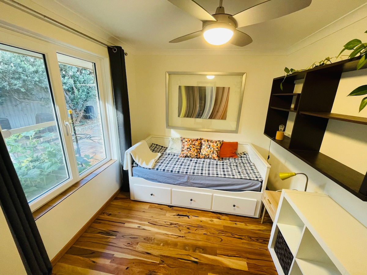 A cozy room features a spacious extendable bed framed by a large window, allowing natural light to fill the space. A colorful arrangement of cushions adds a touch of comfort. A ceiling fan and wooden flooring contribute to a warm, inviting environment.