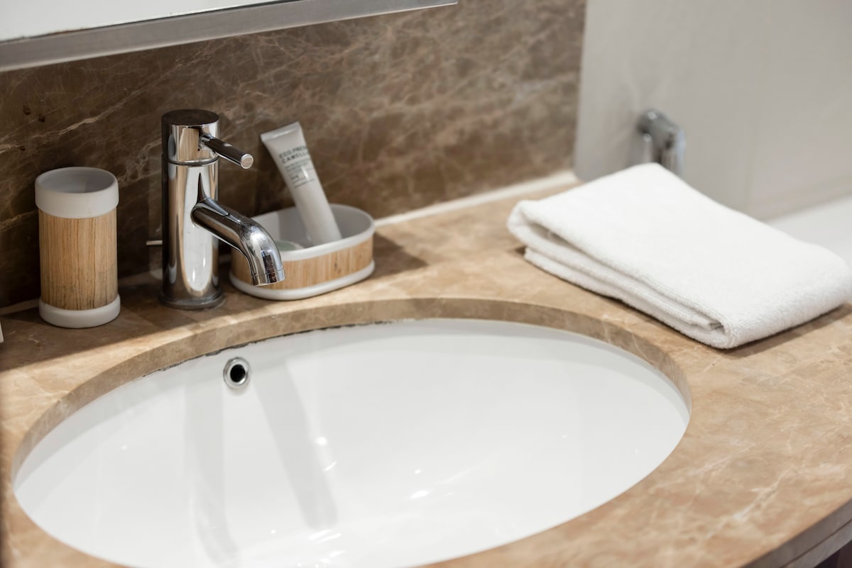 The bathroom sink area features a smooth, oval basin set in a marble countertop. A sleek faucet stands beside neatly rolled white towels, while a small container holds toiletries. Natural elements are suggested by wooden accents in the surrounding holders.