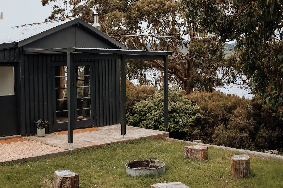 An outdoor space features a cozy fire pit surrounded by wooden stumps on a grassy area. The cabin, designed with a dark exterior, is set against a backdrop of lush trees and views of the river.