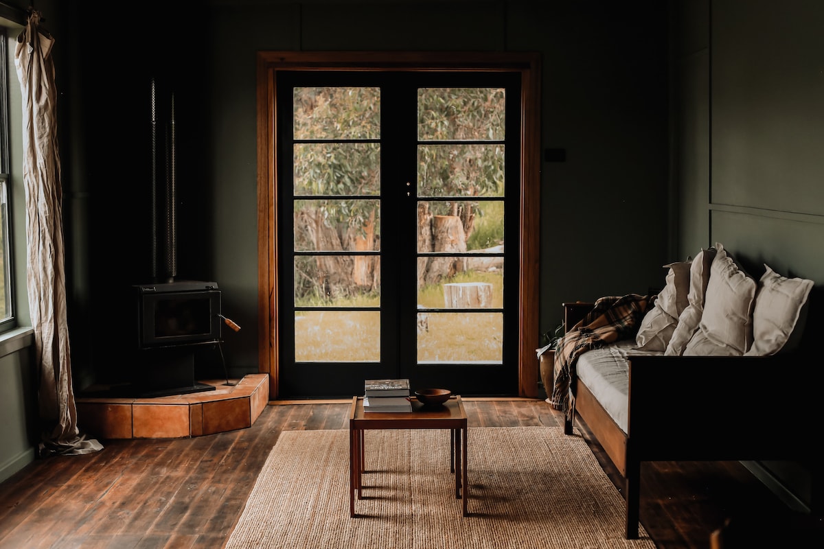 The living area features a comfortable couch adorned with soft pillows, alongside a wooden coffee table. Large glass doors frame the view of the outdoors, allowing natural light to fill the space. A wood heater is positioned on a raised platform, contributing to the cozy ambiance.