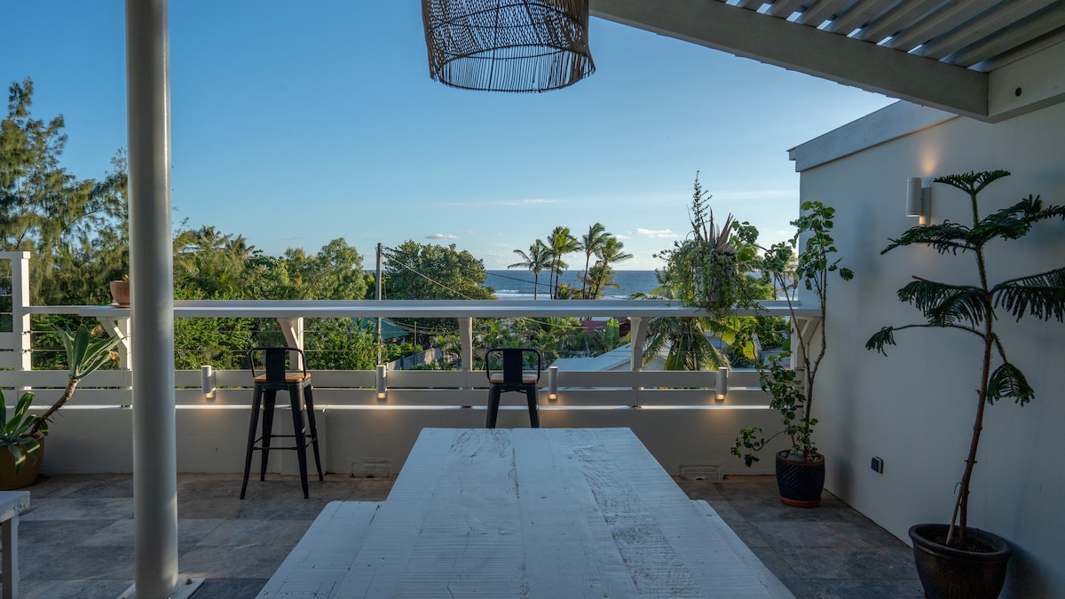 An outdoor terrace is showcased, featuring a long white table surrounded by black stools. Lush greenery can be seen along the railing with palm trees in the background, providing a view of the ocean on the horizon under a clear blue sky.