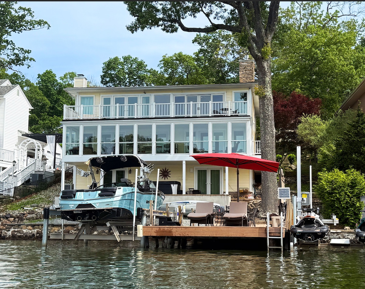 The exterior of a waterfront house is shown, featuring three levels with multiple balconies and large windows. A private dock is visible in front, equipped with seating and a shaded area with an umbrella. A boat is docked alongside, highlighting access to water activities.