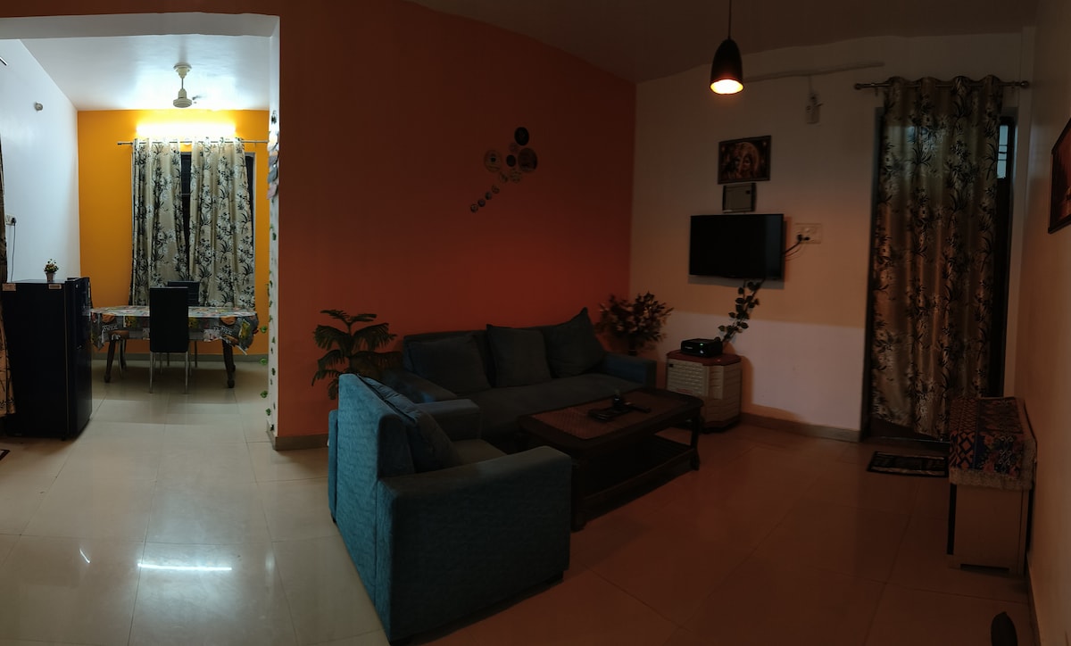 A living room area presents a comfortable seating arrangement with a plush blue sofa. A small coffee table complements the seating. A wall-mounted television is visible, alongside decorative plants. The adjoining dining space features a patterned tablecloth and dark dining chairs, separated by a stylish curtain.