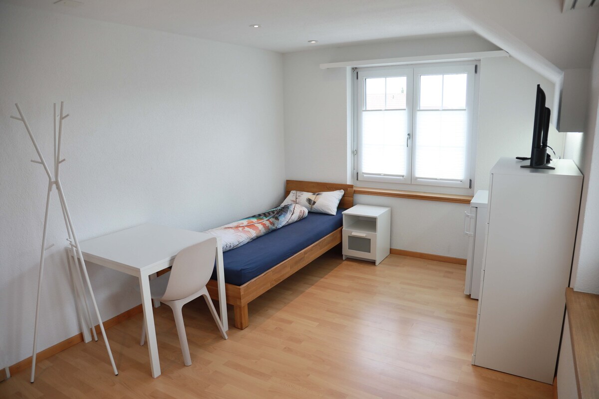 A well-furnished bedroom features a single bed with a colorful blanket, a desk with a chair, and a white storage unit. Large windows provide natural light, and light-colored wooden flooring adds to the room's brightness.