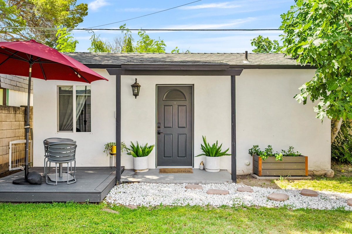 The exterior view of a detached studio guesthouse is shown, featuring a dark front door framed by a covered porch. A small dining set with an umbrella sits on the deck, and a stone path leads to the door. Lush greenery and potted plants add natural elements to the space.
