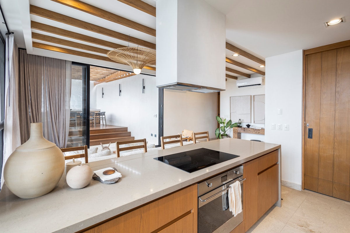 A modern kitchen with wooden cabinetry and a central island is highlighted, featuring an integrated cooktop and minimalist decor. Natural light floods the space through large sliding doors, while wood beams and woven accents add texture to the ceiling. A glimpse of the dining area can be seen beyond.