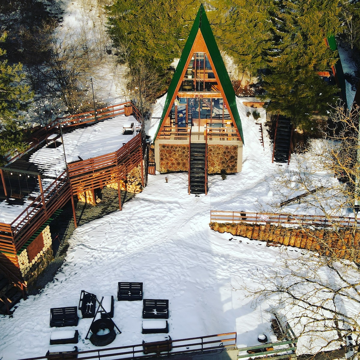 An A-frame cabin is centered in a snowy landscape, surrounded by trees. A multi-level deck with outdoor seating is visible, along with a fire pit area arranged with benches. Snow covers the ground, adding a serene touch to the natural setting.