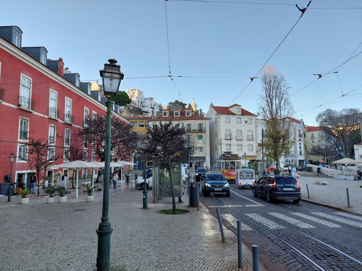 A vibrant street scene is shown, featuring a mix of buildings in various colors including red and white. Paved with cobblestones, the area includes outdoor seating at cafes, parked cars, and tram tracks, with surrounding trees and hills visible in the background.