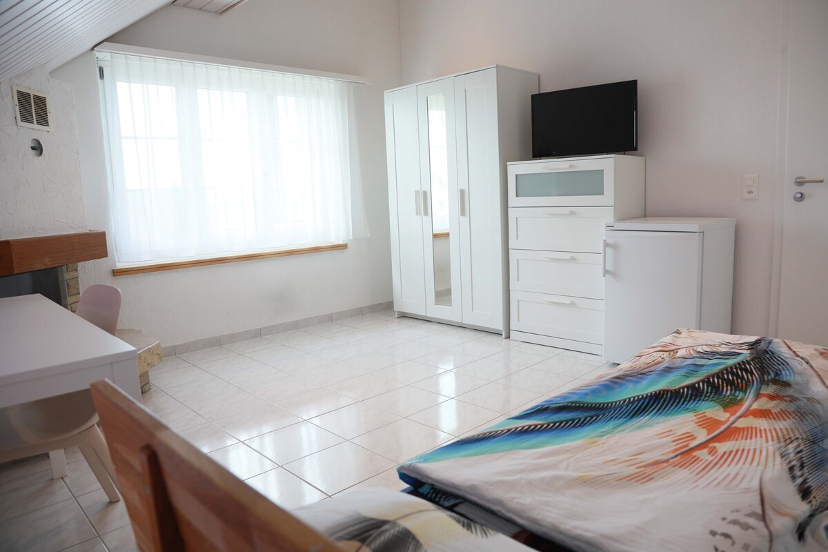 A bright room is characterized by large windows with sheer white curtains letting in natural light. A spacious bed is adorned with a colorful patterned blanket. A white wardrobe, a chest of drawers, and a small refrigerator are neatly arranged, offering practical storage solutions.