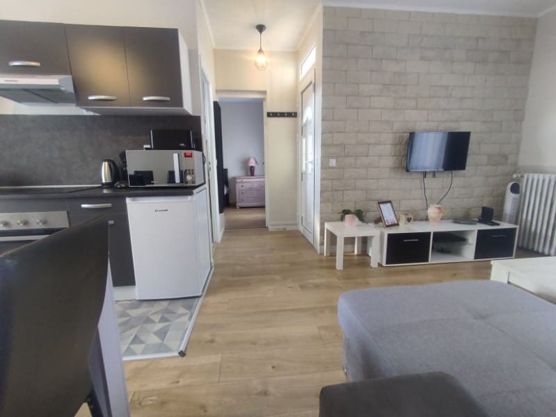 An open living area is displayed, featuring a kitchen with modern appliances and ample countertop space. A comfortable sofa is positioned in front of a wall-mounted television. Light wooden flooring extends throughout, complemented by neutral wall colors and decorative elements on white tables.